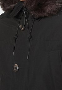 Černá parka s límcem z umělé kožešiny, kovovými cvokovými knoflíky, nastavitelnými šňůrkami a bočními kapsami pro praktičnost.