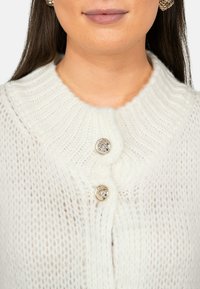 Witte gebreide cardigan met een ribbelkraag, voorzien van twee metalen knopen aan de voorkant. Zachte, fuzzy textuur voegt visuele interesse toe.