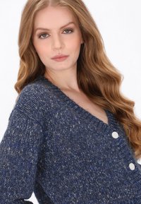 Tmavě modrý strukturovaný kardigan s výstřihem do V, dvěma bílými knoflíky a volným střihem. Smíšená příze s jemnými světle modrými flíčky po celé ploše.