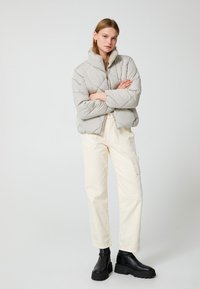 Mujer con una chaqueta acolchada gris claro, pantalones cargo crema y botas gruesas negras, de pie con los brazos cruzados sobre un fondo simple.