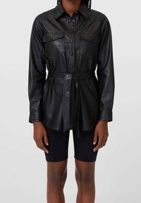 Chemise en cuir noir avec un devant boutonné, deux poches poitrines, une taille ceinturée et des manches longues. Présente un col classique et une texture lisse.