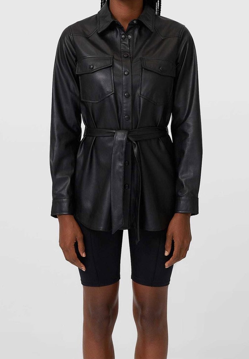 Chemise en cuir noir avec un devant boutonné, deux poches poitrines, une taille ceinturée et des manches longues. Présente un col classique et une texture lisse.