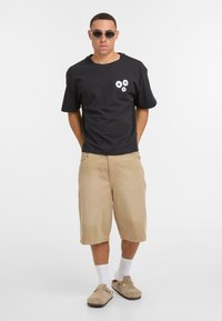 Zwarte katoenen T-shirt met een bloemenontwerp op de borst, beige knielange shorts, witte sokken en beige instapschoenen. Ronde zonnebril.