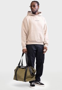 Beige Kapuzenpullover mit vorderer Tasche, schwarze Cargo-Hosen, trägt eine olivgrüne Sporttasche mit schwarzen Riemen und dem Text "rebel". Schwarze Sneaker.