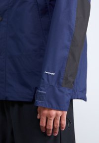 Tamsiai mėlyna striukė su juodais akcentais, ant rankovės pažymėta „DRYVENT“ etiketė, Velcro dirželis ant rankogalių ir lygi audinio tekstūra.