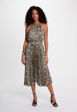 Vrouw met krullend haar, gekleed in een mouwloze beige geplooide jurk met zwarte bloemmotieven en zwarte hooggehakte sandalen met bandjes.