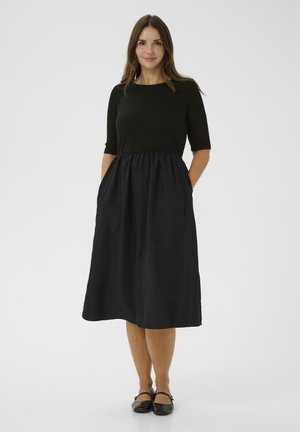 Kvinde iført en sort midi-kjole med albuelange ærmer, hænderne i lommerne, iført sorte flade sko, foran en ensfarvet baggrund.