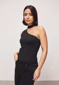 Femme aux cheveux foncés mi-longs portant un top noir asymétrique et un pantalon noir, debout avec une main dans la poche devant un fond uni.