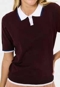 Burgundská pletená polo košile s krátkými rukávy. Obsahuje bílý límec s modrými akcenty a žebrovaný lem a manžety. Knoflíky u výstřihu.