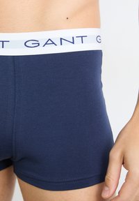Homme portant un boxer bleu marine avec une ceinture élastique blanche arborant le logo GANT bleu marine, main reposant sur le côté, cadré à la taille et à la cuisse.