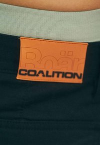 Czarny pasek z jasnoszarym elastanem, z pomarańczową skórzaną naszywką z wytłoczonym logo "Roar COALITION".