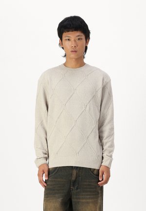 Hellbeige gestrickter Pullover mit einem strukturierten Rautenmuster, geripptem Rundhalsausschnitt und langen Ärmeln, kombiniert mit dunklen, verwaschenen Jeans.