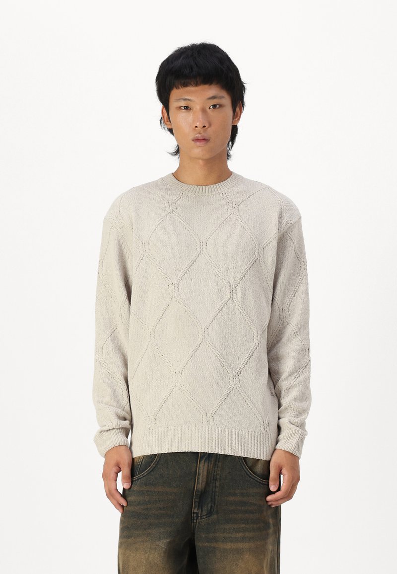 Light beige knit sweater featuring a textured diamond pattern, ribbed crew neckline, and long sleeves, paired with dark, faded jeans.