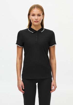 Vrouw draagt een zwart poloshirt met witte bies aan de kraag en mouwen, staand met het gezicht naar voren tegen een effen witte achtergrond.