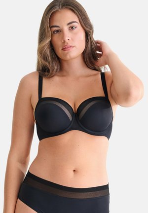 Vrouw met lang bruin haar die een zwarte beugelbeha en bijpassende zwarte slip draagt, poserend met één hand bij haar hoofd.