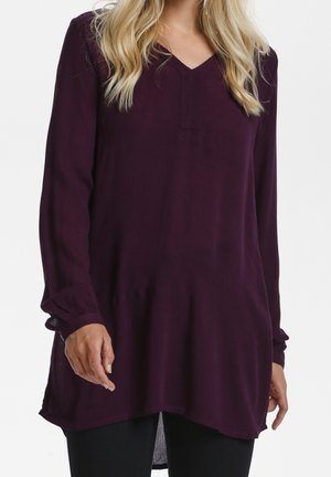 Femme portant un haut tunique à manches longues violet foncé avec col en V et poignets larges, associé à un pantalon noir, vue des épaules jusqu'aux hanches.