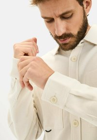 Lys beige knap-op skjorte med et tekstureret stof, som har en synlig lomme, sort accent og store knapper på ærmerne.