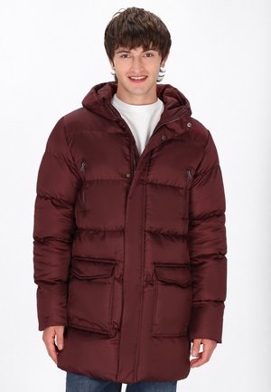 Bordeauxkleurige pufferjack met een capuchon, voorzien van rits- en klepzakken, gestikte textuur en drukknopen, gedragen over een wit shirt.