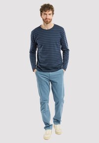 Camicia a maniche lunghe a righe blu navy con colletto rotondo, abbinata a pantaloni azzurri chiari e sneakers beige, in piedi davanti a uno sfondo neutro.