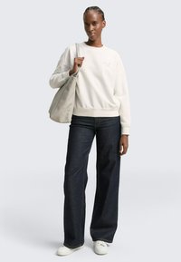 Witte sweater met ronde hals en geribbelde structuur, losvallend, gecombineerd met donkerblauwe jeans met brede pijpen en witte sneakers; draagt een lichtgrijze boodschappentas.