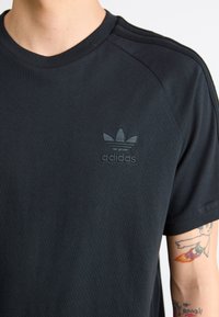 Czarna bawełniana koszulka z ribbowanym okrągłym dekoltem, trzema paskami na ramionach oraz wytłoczonym logo Adidas na piersi.