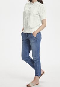 Blusa bianca con texture, maniche a sbuffo corte e chiusura con bottoni, abbinata a jeans blu slim fit con dettagli decorativi sulle tasche.