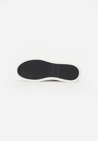 Semelle de chaussure en caoutchouc noir avec un motif texturé. Elle présente un bord blanc et le nom de la marque "Calvin Klein" embossé sur le côté.