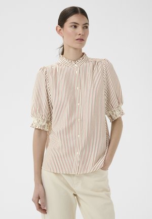 Frau trägt eine cremefarbene Bluse mit roten vertikalen Streifen, ballonförmige kurze Ärmel mit gerüschten Bündchen und beige Hose, stehend mit einer Hand in der Tasche.