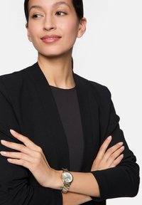 Silberne und goldene Armbanduhr mit rundem Zifferblatt, metallischem Armband und minimalistischem Design, getragen von einer Person mit verschränkten Armen in einem schwarzen Blazer.