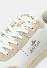 Gros plan sur une basket blanche en cuir avec des accents en suède beige, des lacets blancs et un logo doré "REPLAY" sur le côté et la languette.