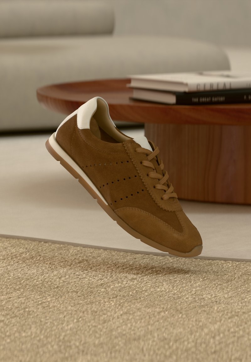 Baskets en daim marron avec un détail blanc au talon flottant au-dessus d'un tapis beige texturé, avec une table ronde en bois et des livres empilés dans une pièce neutre.