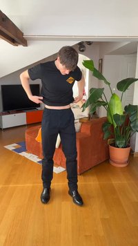 Junger Mann in schwarzem Hemd und Hose hebt sein Hemd, während er in einem Wohnzimmer mit orangefarbenem Sofa, Topfpflanze und Fernseher steht.