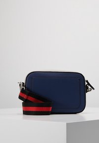 Náměsíčná modrá crossbody taška s texturovaným povrchem, stříbrnými komponenty a černým popruhem se červenými a černými pruhy. Obdélníkový tvar.