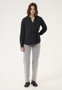 Camicia nera avvolta con chiusure a bottone, maniche lunghe e dettaglio di nodo laterale. Abbinata a jeans dritti grigio chiaro e scarpe nere.