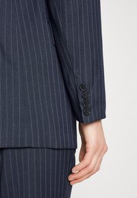 Gros plan sur une main reposant à côté de la manche d'une veste de costume bleu marine à rayures fines, avec quatre boutons noirs, et d'un pantalon assorti.