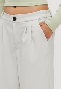 Light grey trousers with a smooth texture, featuring front pleats, a black button closure, and an elastic waistband for comfort.