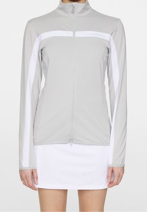 Vrouw draagt een lichtgrijs jack met ritssluiting en witte strepen op de mouwen en borst, gecombineerd met een witte rok.