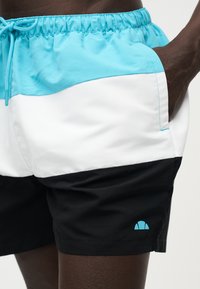 Άνδρας που φοράει μαγιό με μπλοκ χρωμάτων σε μπλε, άσπρα και μαύρα τμήματα, με το χέρι στην τσέπη και μικρό μπλε λογότυπο στο μαύρο τμήμα.