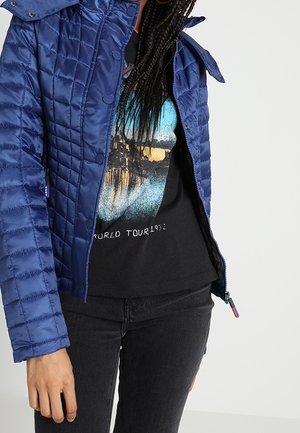 Personne portant une veste matelassée bleue par-dessus un t-shirt graphique noir avec le texte "World Tour 1972", associée à un jean noir.