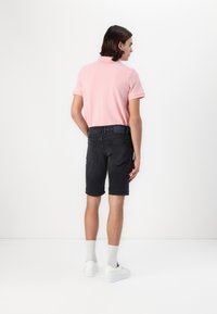 Polo shirt rose clair, shorts en denim noir avec ourlets retournés, et baskets blanches ; présentés de dos sur un fond uni.