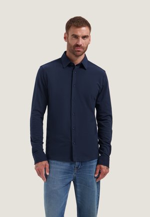 Man die een donkerblauw overhemd met knopen en lange mouwen en lichtblauwe spijkerbroek draagt, staand tegen een effen achtergrond.