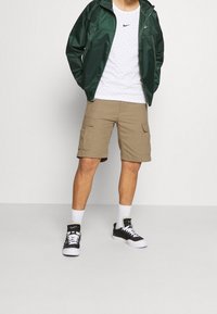 Veste légère verte, t-shirt à motifs blancs, shorts cargo beiges et baskets noires avec semelles et accents blancs. Style sportif.