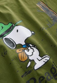 Vihreä oliivinvärinen T-paita, jossa on graafinen kuva sarjakuvakoirasta, joka pitää päässään vihreää hattua ja kantaa ruskeaa laukkua. Taustalla on painettu puu.