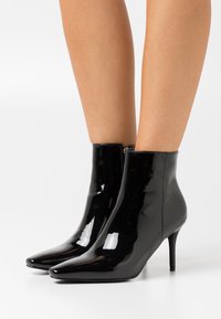 Bottes basses en cuir verni noir avec bout pointu et talon aiguilles fin, présentant une texture lisse et brillante ainsi qu'une fermeture éclair sur le côté.