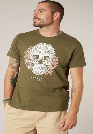 Homme portant un t-shirt vert olive avec un design de crâne décoratif et des roses roses, un pantalon beige et des tatouages visibles sur le bras, regardant vers la gauche.