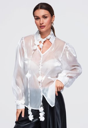 Femme portant un chemisier blanc transparent avec des décorations florales et un pantalon en cuir noir, posant avec la main sur la hanche sur un fond gris.