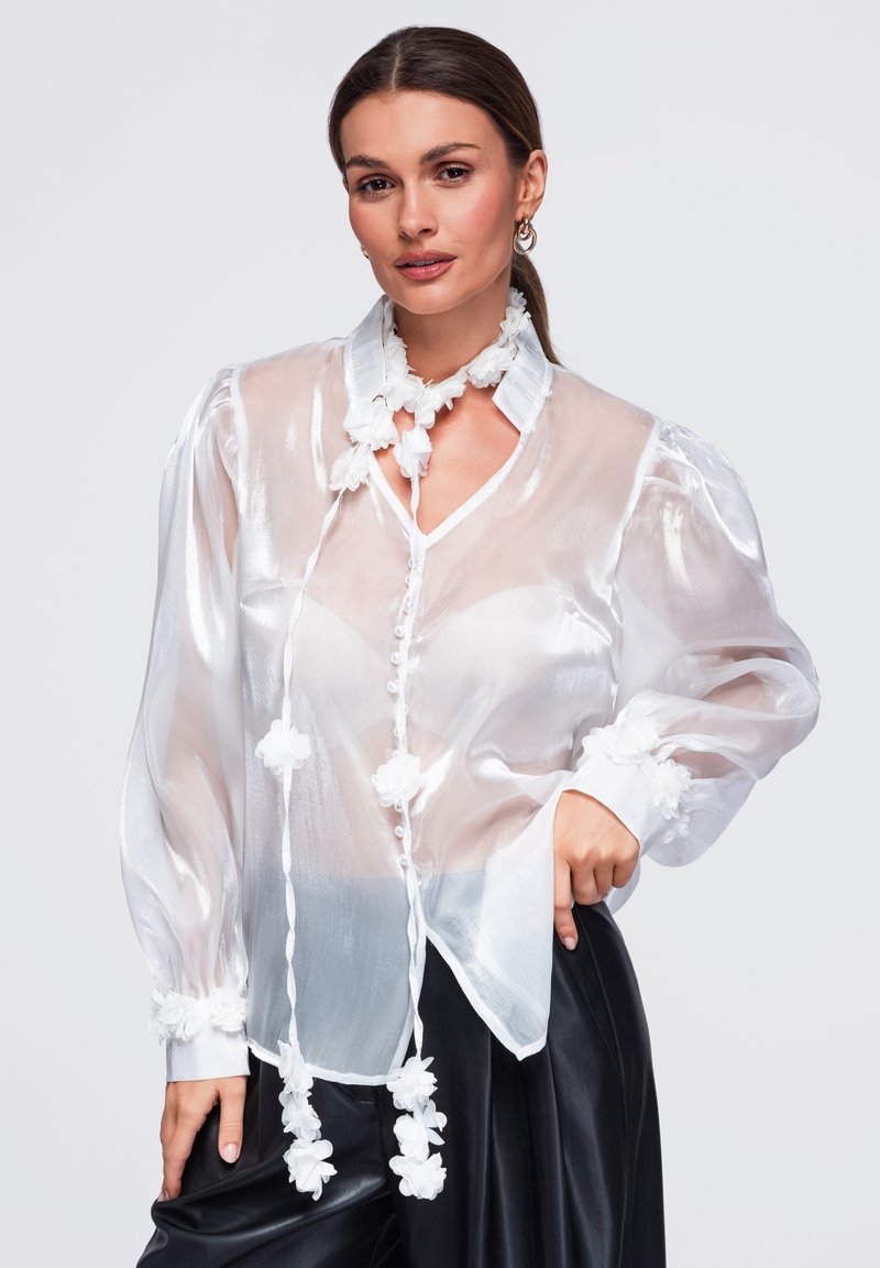 Femme portant un chemisier blanc transparent avec des décorations florales et un pantalon en cuir noir, posant avec la main sur la hanche sur un fond gris.