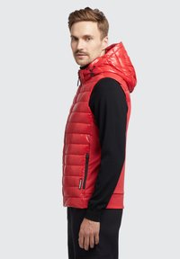 Rød puffervest med hætte, horisontalt quiltet design, lynlåslomme på siden og sort sweatstof på ærmerne og ryggen.