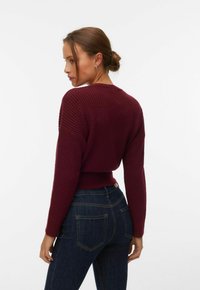 Femme aux cheveux bruns attachés en chignon, portant des boucles d'oreilles, un pull côtelé bordeaux et un jean taille haute bleu foncé, légèrement de profil.