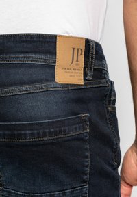 Jeans in denim blu scuro con una patch in pelle che riporta il testo "JP 1880". I jeans presentano cuciture visibili e un design classico delle tasche.